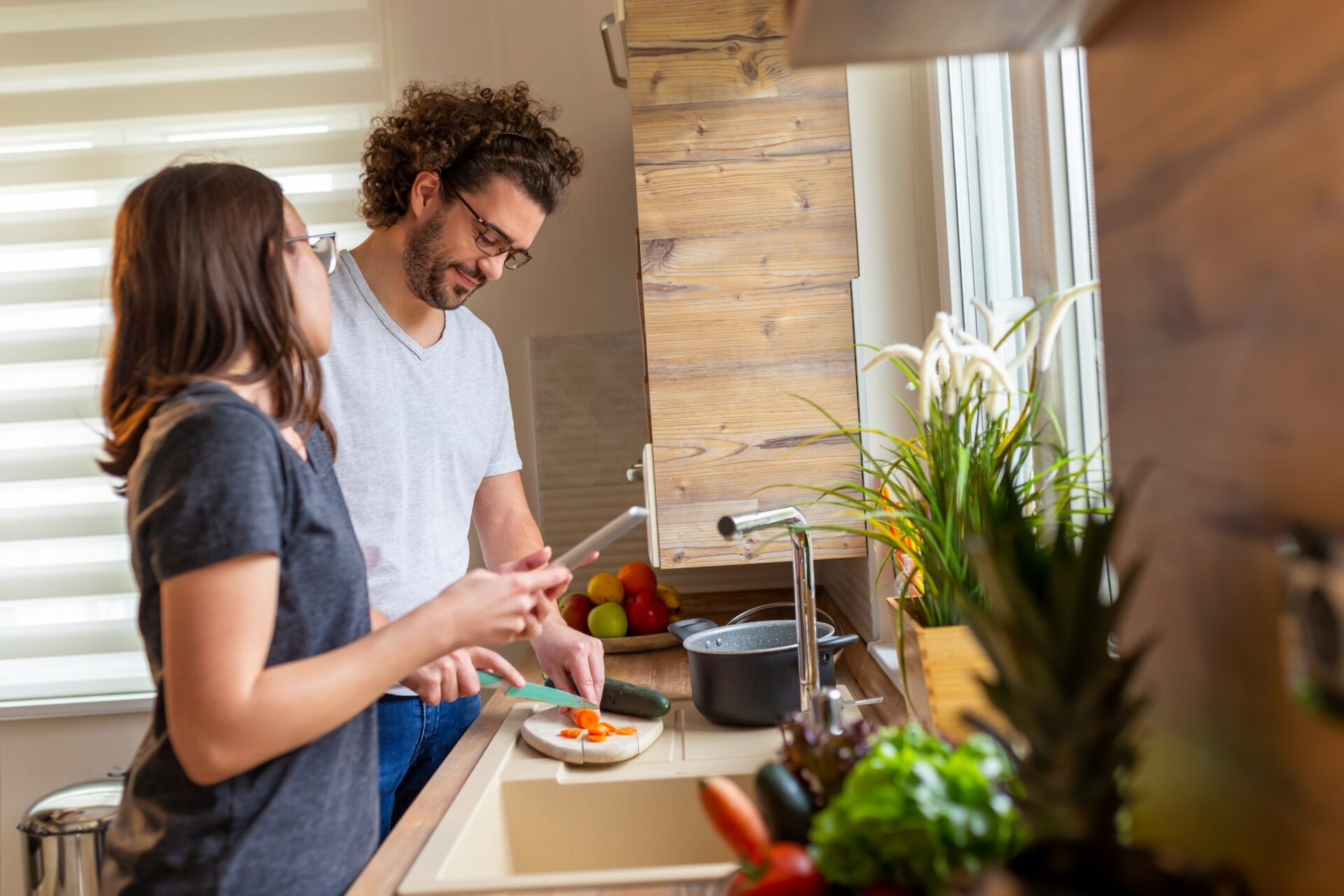 Couple s'amuse dans la cuisine tout en cuisinant ensemble, l'homme coupant des légumes pendant que la femme lit une recette à l'aide d'une tablette