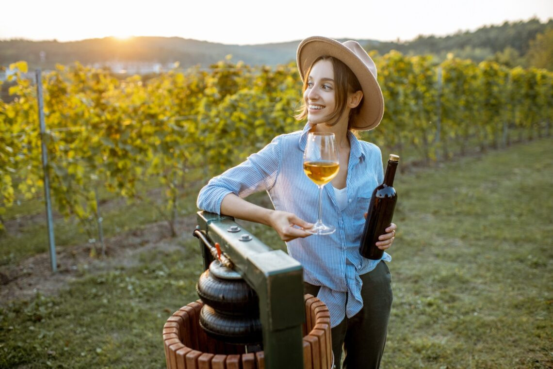 Femme jeune et joyeuse a goûté au vin, tout en se tenant près de la machine de presse sur le vignoble lors d'une soirée ensoleillée